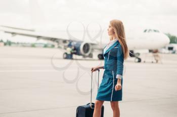 Sexy stewardess with suitcase on aircraft parking, tail of the airplane on background. Air hostess in suit with luggage, flight attendant occupation, aviatransportations job