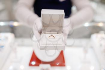 Female seller hands holds gold diamond ring in a box, jewelry store. Salewoman at the showcase holds decorative case with golden decoration, jewellery shop
