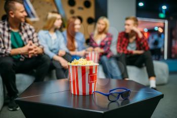 Group of teenagers relax on the couch and waiting for showtime in cinema hall. Male and female youth sitting on sofa in movie theater, popcorn on the table
