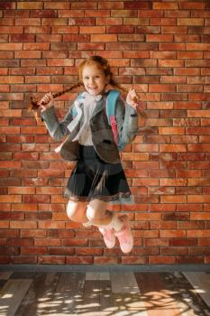 Cute schoolgirl with schoolbag jumps at the brick wall. Playful female pupil with backpack poses in the school
