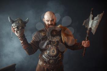 Angry viking dressed in traditional nordic clothes holds enemy's skull in helmet and axe, barbarian image, murderer. Ancient warrior in smoke