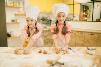 Two smiling little girls cooks in caps and aprons having fun, cookies preparation on the kitchen. Kids cooking pastry, children chefs preparing cake