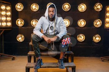 Black rapper in hoodie sitting on the steps, perfomance on stage with spotlights on background. Rap performer on scene with lights, underground music, urban style