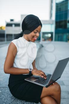 Young business woman uses laptop against office building. Smiling black businesswoman in skirt and white blouse works outdoors