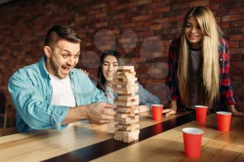 Smiling friends plays table game at home, selective focus on tower. Board game with wooden blocks requiring high concentration, entertainment for funny company