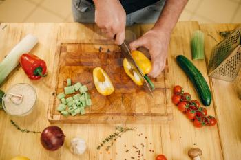 Male chef with knife cuts yellow pepper on wooden board, top view. Man cutting vegetables, fresh salad cooking, kitchen interior on background
