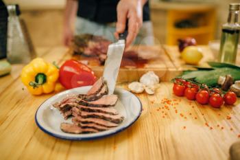 Male chef with knife puts roasted meat slices into the plate, kitchen on background. Man preparing beef with vegetables on countertop