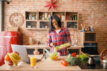 Black woman looking recipe on laptop, young ethnic girl cooking on the kitchen. African female person preparing vegetable salad
