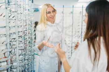 Female optician and customer shooses glasses in optics store. Selection of eyeglasses with professional optometrist. Eye care, spectacles choice