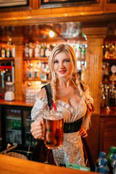 Sexy waitress held out a mug of fresh foamy beer at the counter in pub. Octoberfest barmaid with attractive shapes in traditional style dress