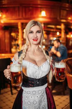 Sexy waitress holds two mugs of fresh beer in pub. Octoberfest barmaid with attractive shapes