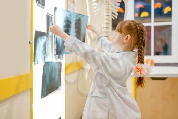 Little girl in uniform looks at the x-ray, playing doctor, playroom. Kid plays medicine worker in imaginary hospital, profession learning