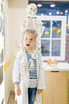 Little girl in uniform near a human skeleton, playing doctor, playroom. Kid plays medicine worker in imaginary hospital, profession learning, childish dream