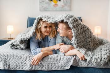 Young love couple lying in bed and peeking out from under the cover. Husband and wife having fun in bedroom. Happy family together