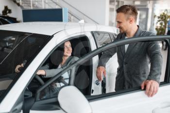 Happy couple buying new car in showroom, woman behind the wheel. Male and female customers choosing vehicle in dealership, automobile sale, auto purchase