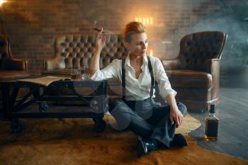 Woman in strict clothes relax on the floor with whiskey and cigar, retro fashion, gangster style. Vintage business lady in office with brick walls