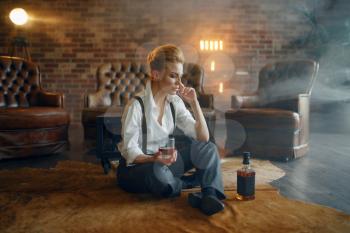 Woman in strict clothes sitting on the floor with whiskey and cigar, retro fashion, gangster style. Vintage business lady in office with brick walls