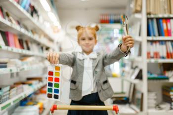 Little school girl with cart, watercolor paints and brushes, shopping in stationery store. Female child buying office supplies in shop, schoolchild in supermarket