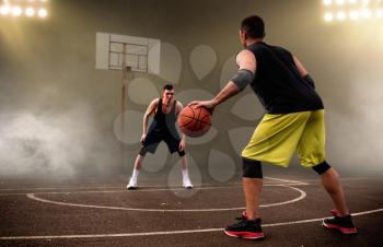 Two basketball players work out tactics, dark background. Male athletes in sportswear play the game on streetball training