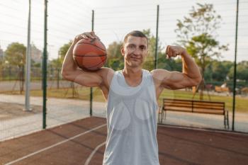 Male basketball player with ball shows his muscles on outdoor court, front view. Male athlete in sportswear on streetball training, summer stadium