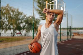 Tired male basketball player with ball on outdoor court, front view. Male athlete in sportswear on streetball training, summer stadium