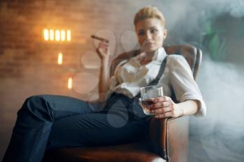 Woman in shirt and trousers sitting in leather chair with whiskey and cigar, retro fashion, gangster style. Vintage business lady in office with brick walls