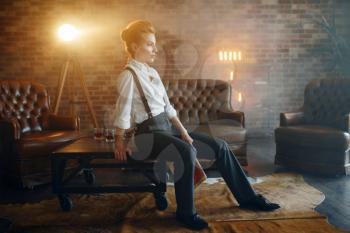 Woman in shirt and trousers with suspenders poses in studio, retro fashion, gangster style. Vintage business lady in office with brick walls