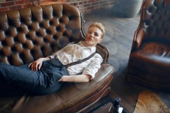 Woman in strict clothes lying on leather couch, retro fashion, gangster style, female macho. Vintage business lady in office with brick walls