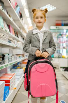 Little schoolgirl with backpack in hand at the shelf in stationery store. Female child buying office supplies in shop, schoolchild in supermarket