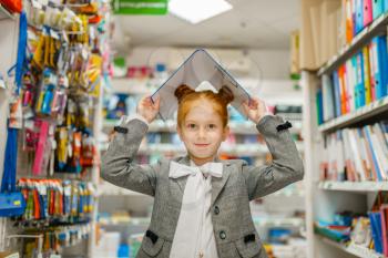 Little schoolgirl holds folders over her head in stationery store. Female child buying office supplies in shop, schoolchild in supermarket