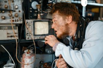 Crazy engineer looks on oscilloscope display in laboratory. Electrical testing tools on background. Lab equipment, engineering workshop
