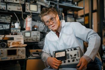 Crazy scientist holds electrical device in laboratory. Electrical testing tools on background. Lab equipment, engineering workshop