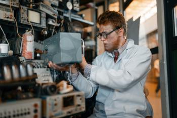 Crazy scientist in glasses holds electrical device in laboratory. Electrical testing tools on background. Lab equipment
