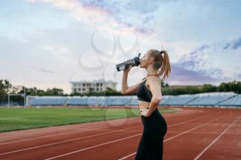 Female runner in sportswear drinks water, training on stadium. Woman doing stretching exercise before running on outdoor arena