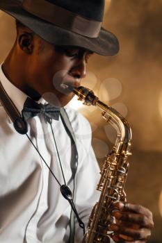 Black jazzman in hat plays the saxophone on the stage with spotlights. Black jazz musician preforming on the scene