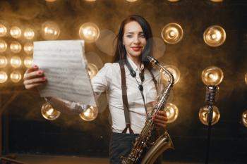 Female saxophonist with saxophone holds music book on the stage with spotlights. Jazz performer playing on the scene