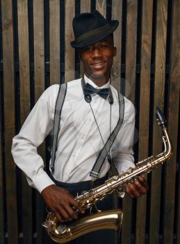Black smiling jazz musician poses with saxophone, wooden fench background. Black jazzman in hat poses with instrument