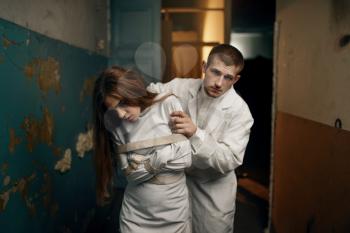 Male psychiatrist leads crazy female patient in straitjacket, mental hospital. Woman in strait jacket undergoing treatment in clinic for the mentally ill