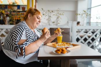 Fat woman eating high calorie food in fastfood restaurant. Overweight female person at the table with junk dinner, obesity problem