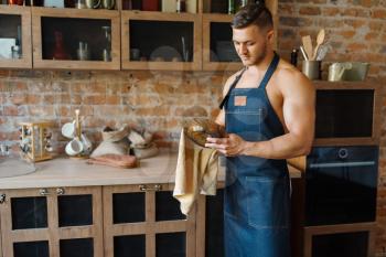 Nude husband in apron wipes the dishes on the kitchen. Naked male person preparing breakfast at home, food preparation without clothes