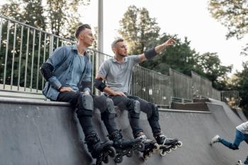 Roller skating, two male skaters sitting on the ramp in park. Urban roller-skating, active extreme sport outdoors