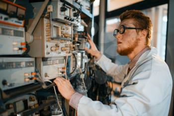 Crazy scientist in glasses adjusts electrical device in laboratory. Electrical testing tools on background. Lab equipment, engineering workshop
