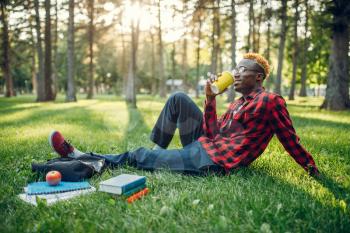 Black student drinks coffee on the grass in summer park. A teenager studying outdoors and having lunch