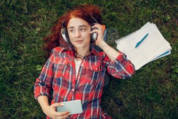 Female student in headphones lying on the grass in summer park. Ginger teenager studying and leisures outdoors