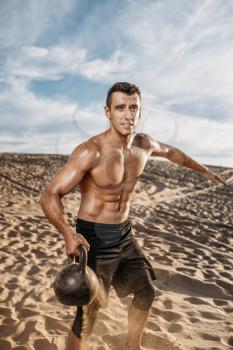 Male athlete doing exercises with kettlebell in desert at sunny day, flying sand effect. Strong motivation in sport, strength outdoor training