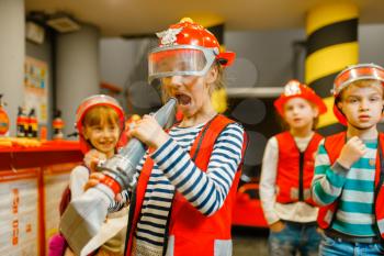 Female child in helmet and uniform having fun with hose in hands playing fireman, playroom indoor. Kids lerning firefighter profession. Children lifeguards, little heroes in equipment on playground
