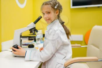 Little girl in uniform sitting at the microscope and playing doctor, playroom. Kid plays medicine worker in imaginary hospital lab, profession learning, childish dream