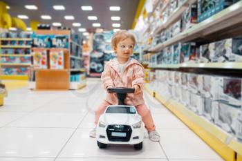 Little girl on electromobile at the shelf in kids store, front view. Daughter choosing toys in supermarket, family shopping, young customer