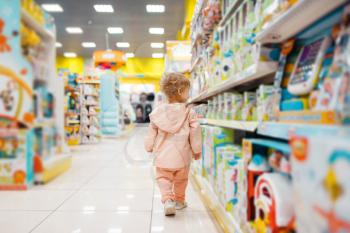 Little girl at the shelf choosing toys in kids store, side view. Daughter in supermarket, family shopping, young customer