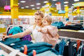 Mother with her little daughter choosing clothes in kids store. Mom and child buying dress in supermarket together, family shopping